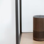 A sleek, modern smart speaker displayed on a wooden shelf in a minimalist room setting.