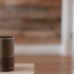A stylish smart speaker sits on a wooden surface in a cozy, warm indoor setting.
